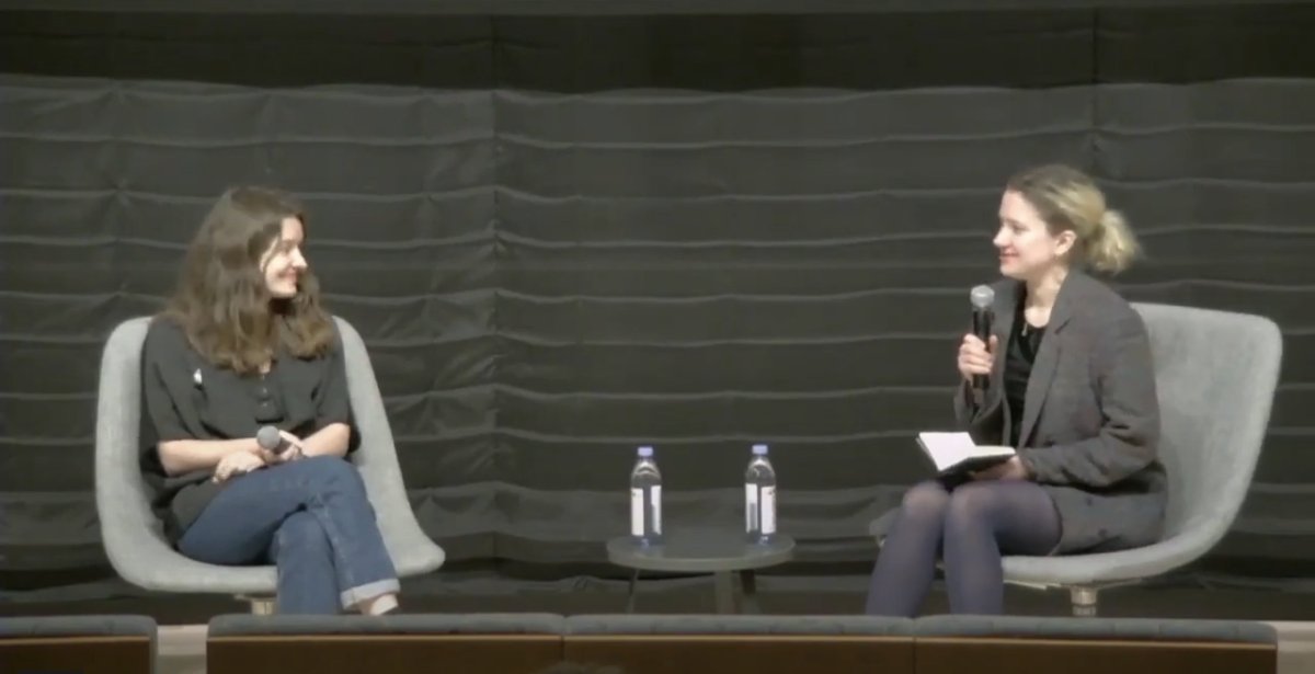 A screenshot from a video of the event, Anna Tropnikova and Ukrainian filmmaker Iryna Tsilyk sit in chair's facing one another on a dark grey stage. The women are smiling and holding microphones.
